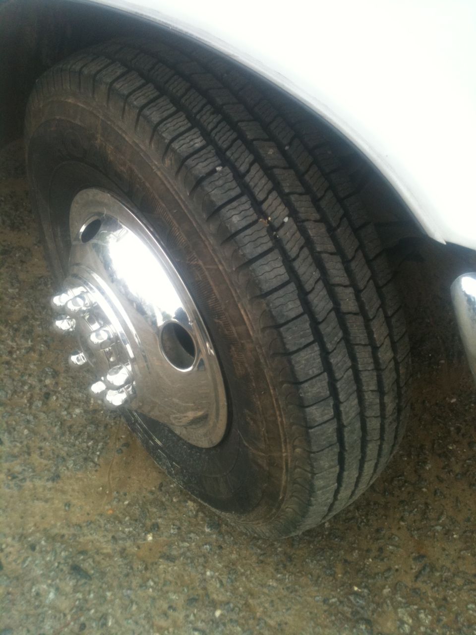White truck wheel with chrome hubcap and tire. Ground is gravel