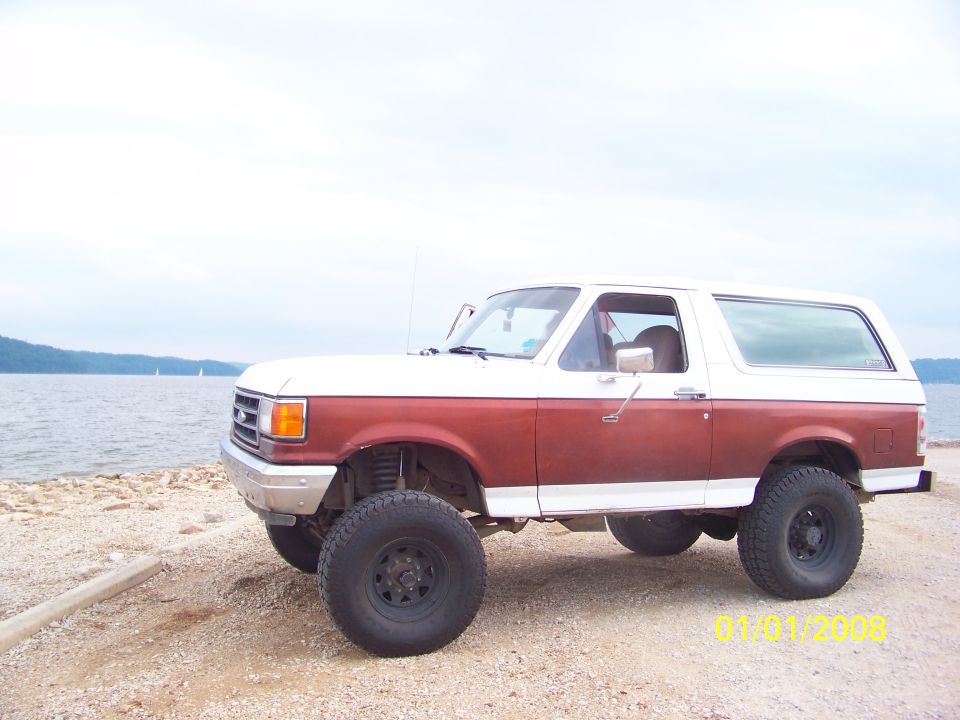1989 Ford Bronco with two-tone paint, lifted suspension, and off-road tires parked on a gravel shore next to a body of water