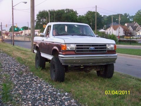 1989 Ford Bronco - This lifted Ford pickup truck, likely from the late 1980s, is parked on a grassy shoulder next to a road.