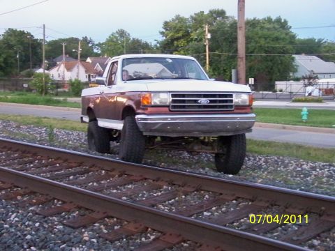 1989 Ford Bronco - A lifted Ford pickup truck is parked on railroad tracks, with houses and trees in the background.