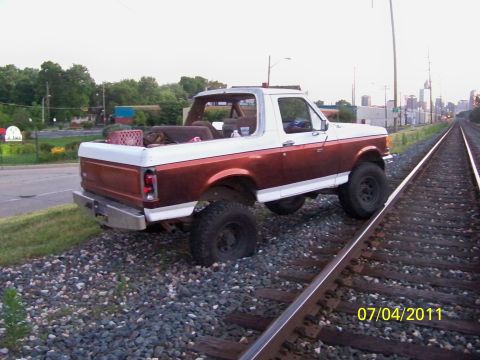 1989 Ford Bronco - Rear three-quarter view of a lifted two-tone brown-and-white Ford Bronco with the hardtop removed, showing the exposed roll bar, rear bench…