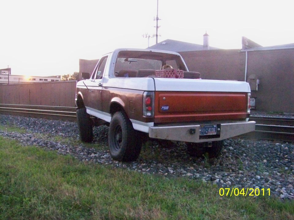 Rear view of a brown and white 1989 Ford Bronco with aftermarket bumper parked by railroad tracks