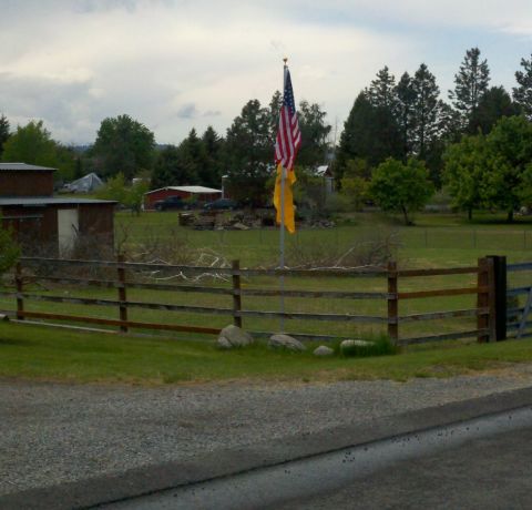 1978 Ford Mustang II - An American flag flies over a rural landscape with a wooden fence and buildings under a cloudy sky.