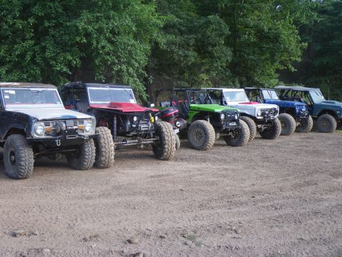 1995 Ford Bronco - Multiple heavily modified Ford Broncos are lined up on a dirt surface with trees in the background.