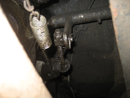 Engine-bay linkage with a coiled return spring and nut-and-bolt adjustment on a greasy black bracket