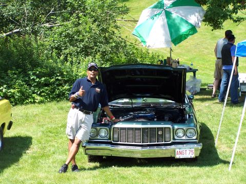 1975 Ford Ranchero - A man poses with a green 1975 Ford Ranchero at an outdoor car show. The vehicle's hood is open, revealing the engine.