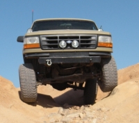 1986 Ford Bronco - This tan Ford pickup truck is shown from the front, navigating a rocky, off-road path.