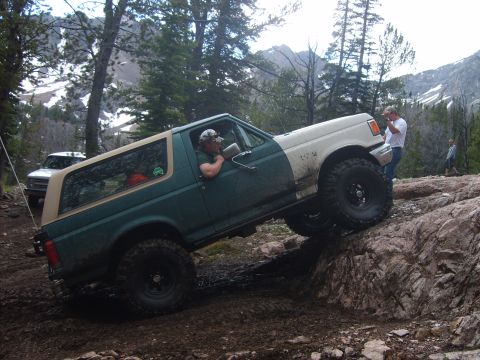 1989 Ford Bronco - A two-tone Ford Bronco navigates a rocky, muddy trail with trees and mountains in the background.