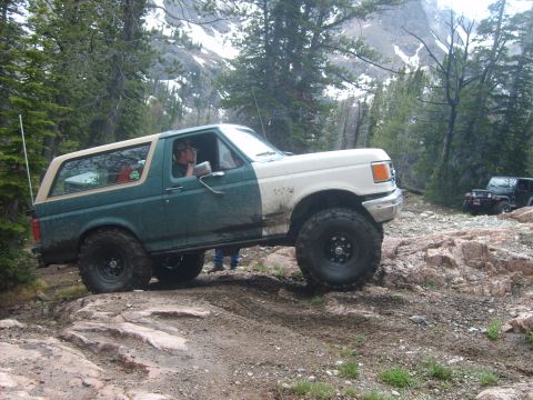 1989 Ford Bronco - A two-tone Ford Bronco tackles a challenging off-road path in a mountainous, wooded area.