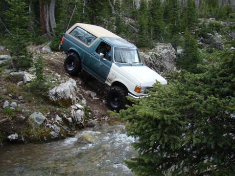 1989 Ford Bronco - A two-tone Ford Bronco navigates a rocky stream bed in a wooded mountain area.