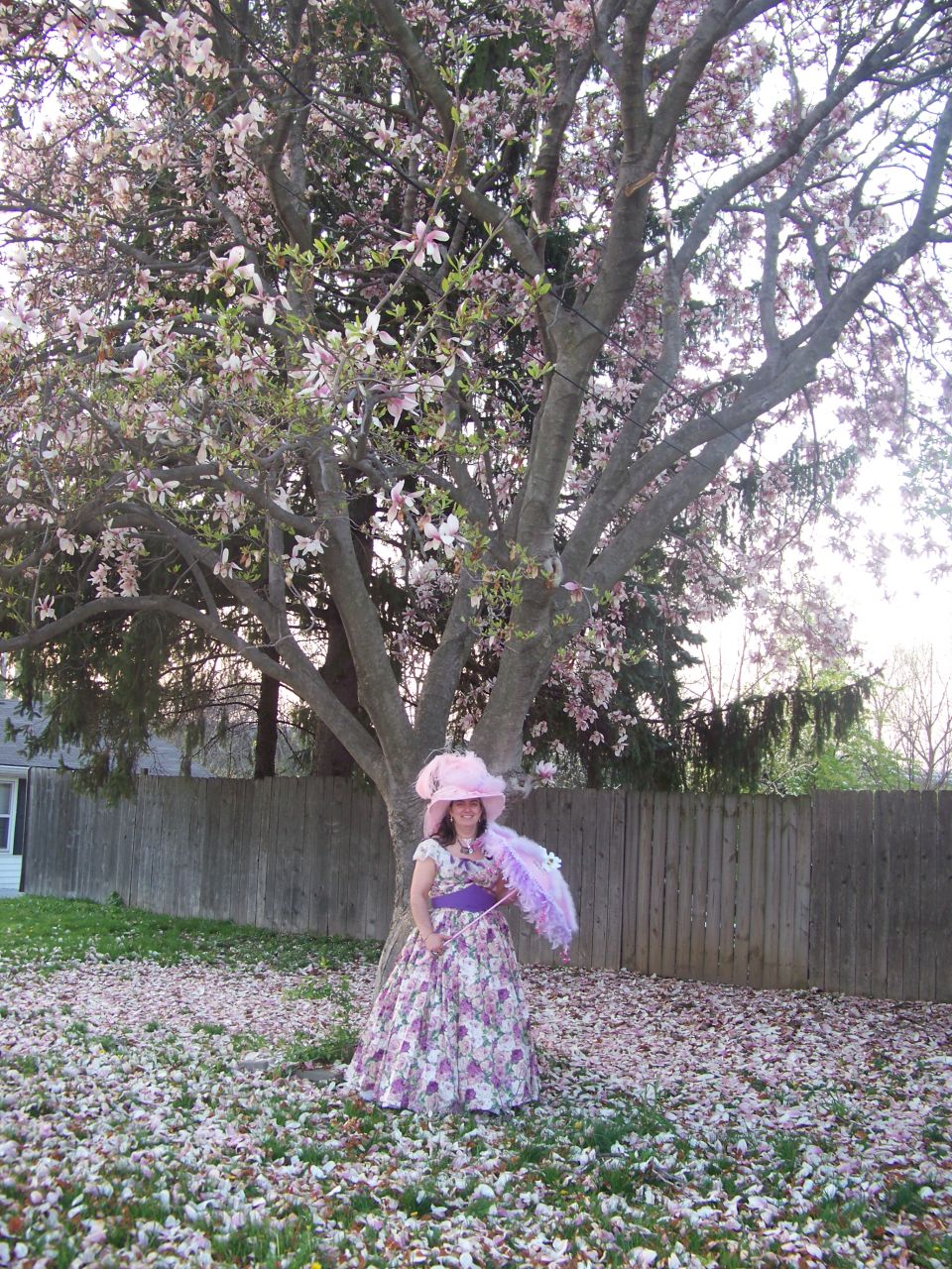 Woman in a long floral dress and large pink hat holding a parasol under a blooming tree
