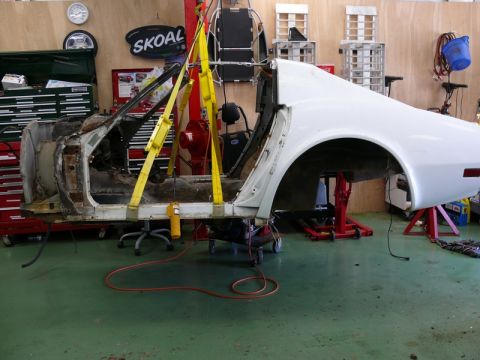 1976 Chevrolet Corvette - A white car body, likely a 1976 Chevrolet Corvette, is suspended by yellow straps and supported by red repair stands during restoration.