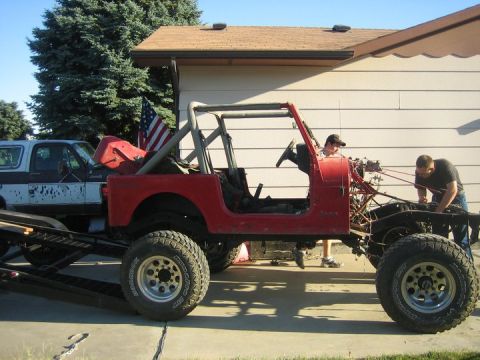1990 Jeep YJ - Two people work on a red Jeep CJ-series, which is on a trailer next to another vehicle.