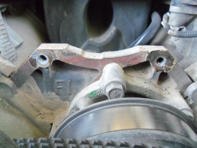 Close-up of a rusty brake caliper bracket with mounting holes and a bolt. A brake rotor and belt are visible below