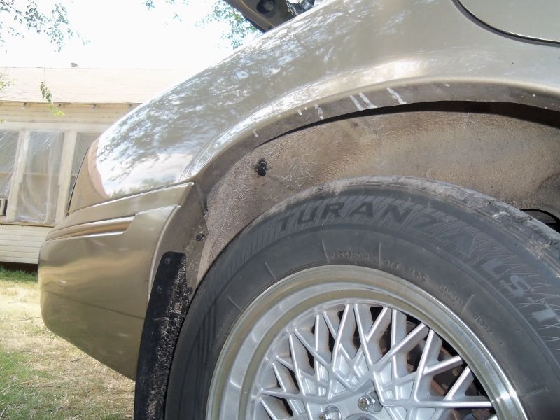 Close-up of a car's rear wheel well, showing the tire and part of the fender. A house is visible in the background