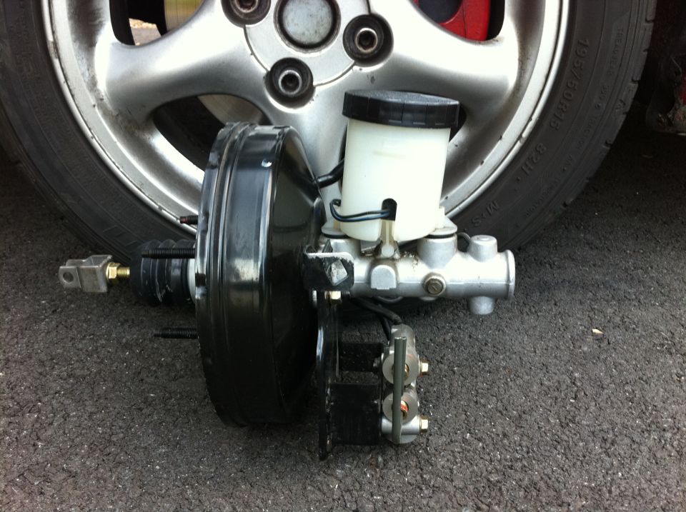 Brake booster and master cylinder assembly on pavement in front of a 195/50R15 alloy wheel