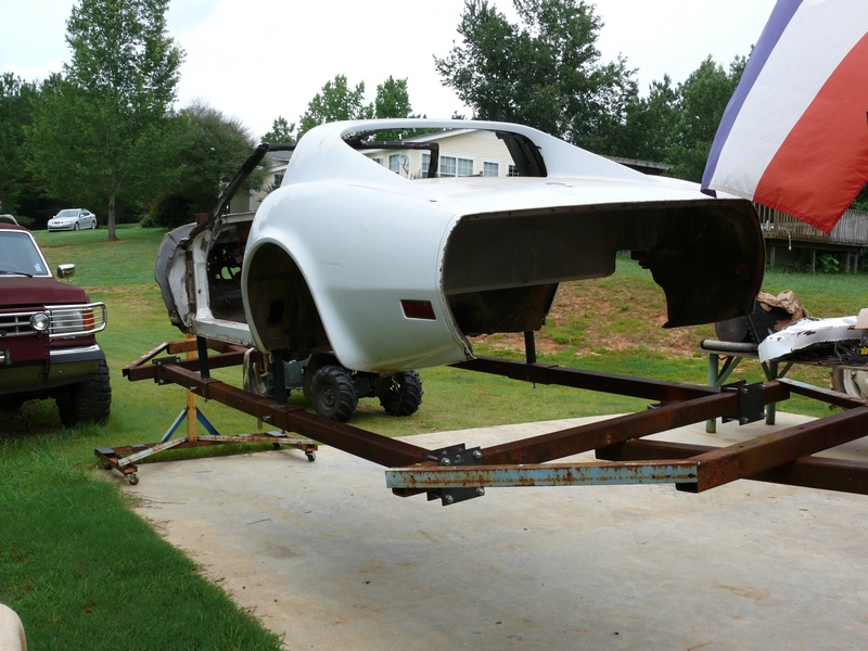 White Corvette body on a trailer, with a red pickup truck and a silver car in the background