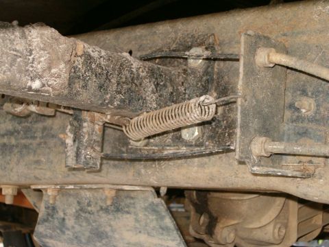 1991 Ford Bronco - Tight underside view of a dirt-caked truck frame rail with a coil return spring stretched between a welded tab and a pivot lever, flanked b…