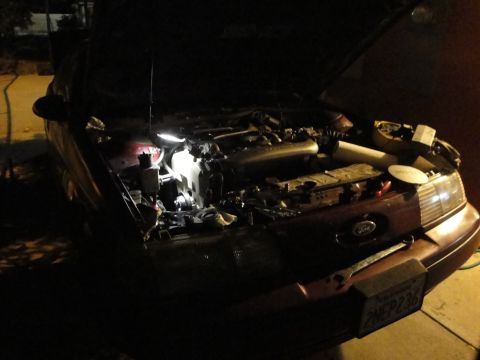 1993 Ford Taurus - The engine bay of a 1993 Ford Taurus is visible with the hood open and a work light illuminating the engine components.