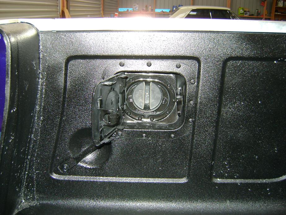 Open fuel door and round gas cap on black bedliner-coated panel of a vehicle in a garage