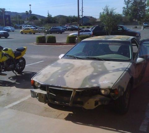 2006 Ford E350 - A car with a camouflage paint job and a damaged front bumper is parked in a lot, with its driver's door ajar.