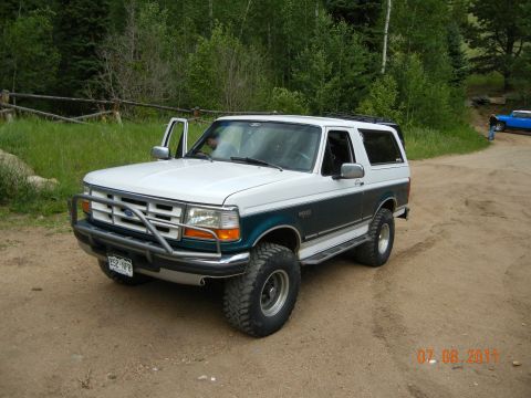 1996 Ford Bronco - A white and teal 1996 Ford Bronco is parked on a dirt road with its driver's door ajar.