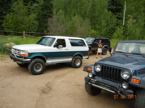 1996 Ford Bronco - Three SUVs, including a 1996 Ford Bronco, are parked on a dirt road with trees in the background.