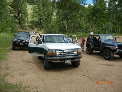1996 Ford Bronco - A group of off-road vehicles are parked on a dirt road in a wooded area.