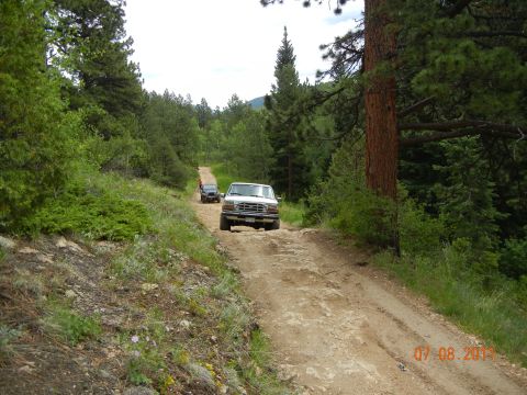 1996 Ford Bronco - Two SUVs navigate a rough dirt road through a forested area.