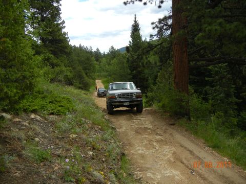 1996 Ford Bronco - Two SUVs navigate a wooded dirt road on an overcast day.