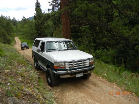 1996 Ford Bronco - A white and teal Ford Bronco navigates a wooded dirt road, with another SUV in the distance.