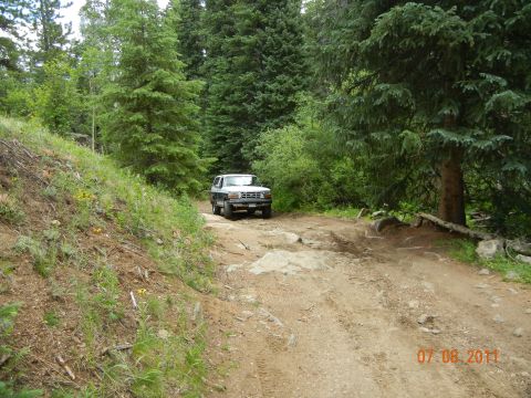 1996 Ford Bronco - A white 1996 Ford Bronco navigates a rough, rocky dirt road through a dense forest.