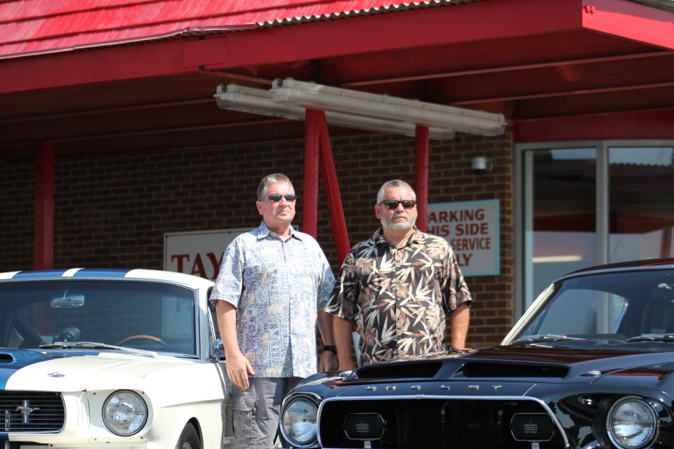Two men stand by a white and blue Mustang and a black Mustang. The black Mustang has 'SUPERMOTORS' logo on the grille
