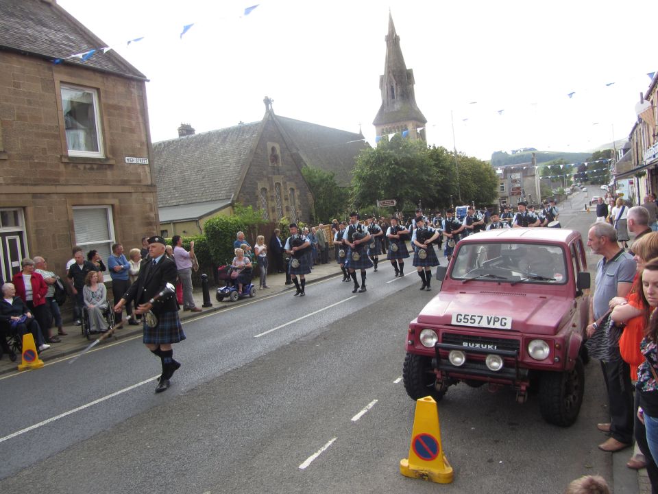Red Suzuki Samurai with license plate G557 VPG driving in a street parade. Bagpipers in kilts march alongside