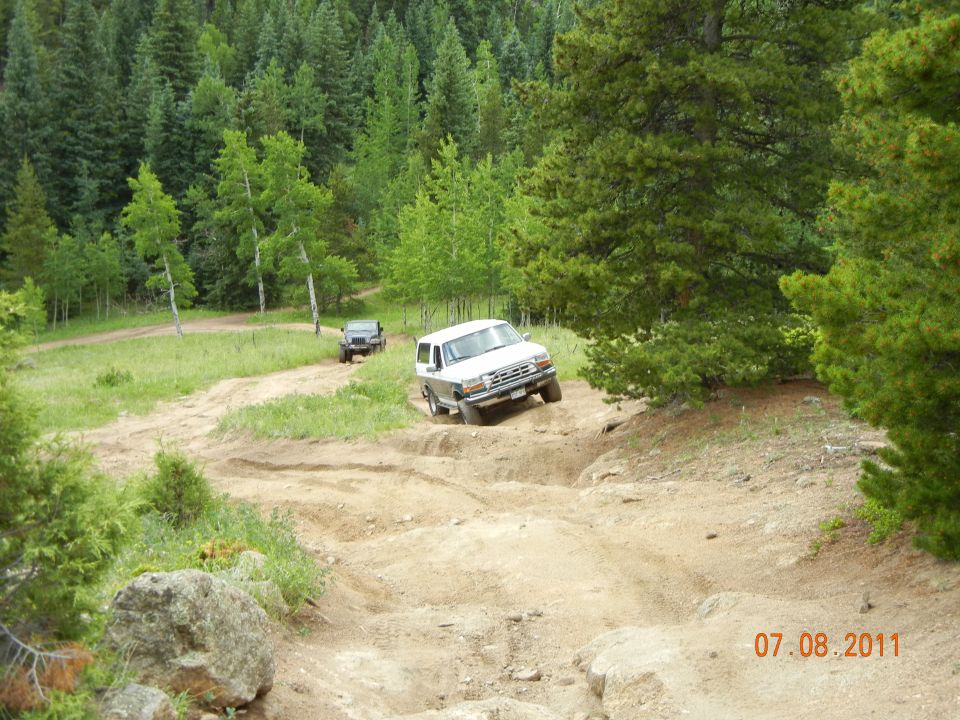 White SUV driving down a steep dirt trail with a dark SUV behind it, surrounded by trees