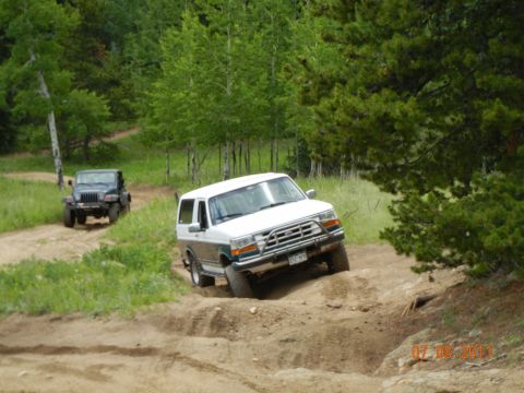 1996 Ford Bronco - Two SUVs navigate a dirt path through a wooded area on a sunny day.