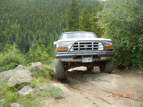 1996 Ford Bronco - 1996 Ford Bronco tackles a challenging off-road obstacle with a forest backdrop.