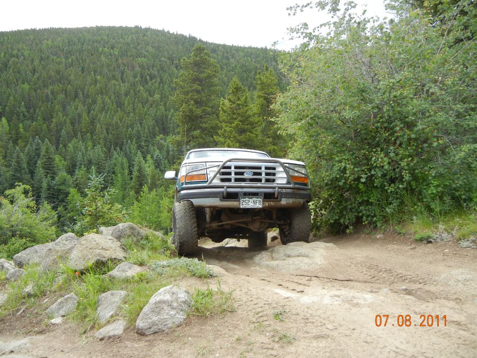 Ford truck with a brush guard driving over rocks on a dirt trail, trees and a hill in the background