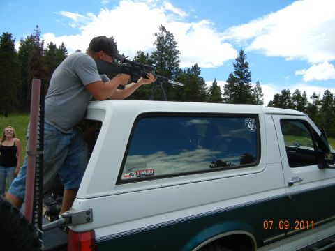 1996 Ford Bronco - A man aims a rifle from the open rear of a white SUV with a green stripe, with a date stamp of 07.09.2011 visible.