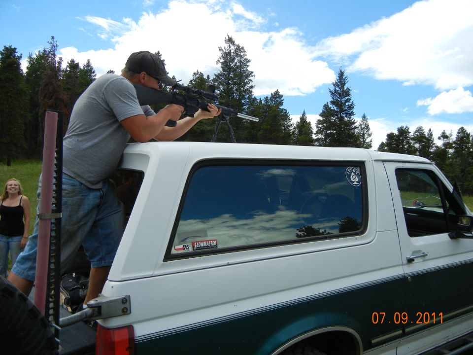 White SUV with green stripe, side view. Man aims rifle from open rear door. Date stamp 07.09.2011