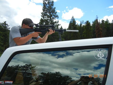 1996 Ford Bronco - A man aims a rifle from the interior of a white truck, with clouds and trees reflected in the window.