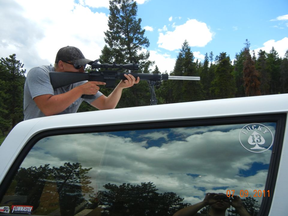 Man aiming rifle from inside white truck cab, reflecting clouds and trees
