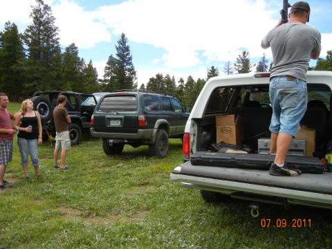 1996 Ford Bronco - A group of SUVs and people are gathered in a grassy field with trees under a cloudy sky.