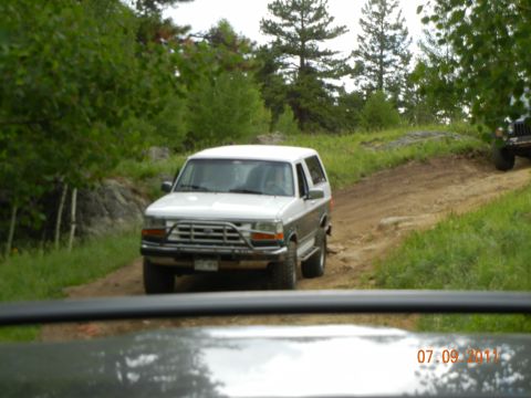 1996 Ford Bronco - White Ford Bronco navigates a dirt path in a wooded area, with another vehicle visible ahead.