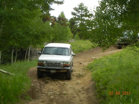 1996 Ford Bronco - A white SUV navigates a dirt path through a wooded area, with another vehicle in the distance.