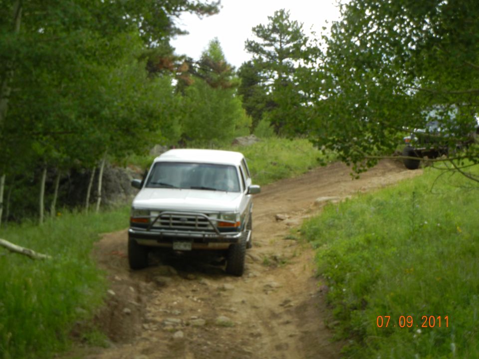 White SUV driving on a dirt road with trees on either side. Another vehicle is visible in the background