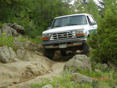 1996 Ford Bronco - A white 1996 Ford Bronco navigates a rocky off-road trail surrounded by trees and vegetation.