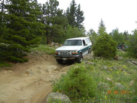 1996 Ford Bronco - A white and green Ford Bronco navigates a challenging off-road path with another vehicle visible in the background.