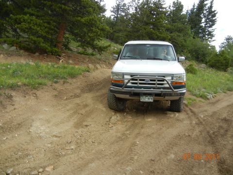 1996 Ford Bronco - This white Ford SUV is navigating a rough dirt road surrounded by trees and vegetation.