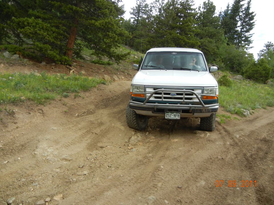 White Ford SUV with brush guard driving on a dirt road in a wooded area. License plate reads 252-NFB